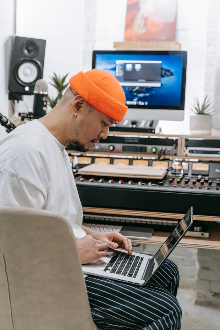 About Asian man wearing orange beanie using laptop in modern home music studio.